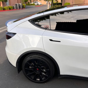 May include: White Tesla Model Y with black wheels and a black rear spoiler. The car has tinted windows and a black door handle.