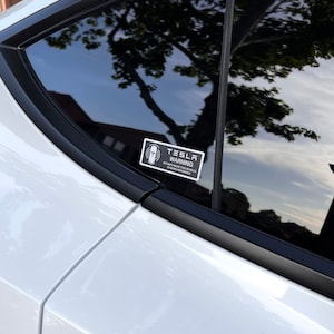 May include: A black and white sticker with the text "Tesla Warning" and a symbol of a person with a circle around them. The sticker is on the back window of a white car.
