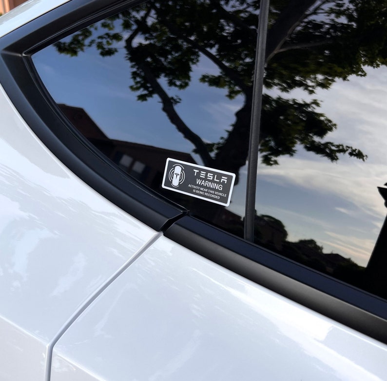 May include: A black and white sticker with the text "Tesla Warning" and a symbol of a person with a circle around them. The sticker is on the back window of a white car.