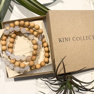 May include: Three bracelets in a box. The bracelets are made of wood beads and white agate stones. The box is brown and has the text "KINI COLLECTIVE" printed on it.