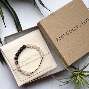 May include: A black lava rock and white agate beaded bracelet with a gold bar accent. The bracelet is packaged in a brown box with the text "KINI COLLECTIVE" printed on the top.