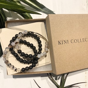 May include: Three bracelets in a brown box. The bracelets are made of black lava rock beads and white agate beads. The box is labeled "KINI COLLECTIVE".