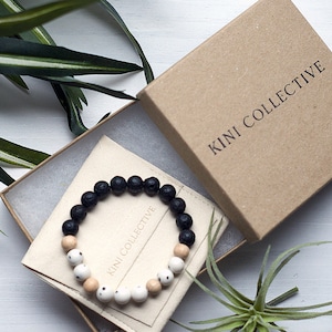 May include: A black and white beaded bracelet with the text "KINI COLLECTIVE" on a white card inside a brown box.
