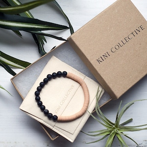May include: A black lava rock and beige beaded bracelet in a small, white box with the text "KINI COLLECTIVE" on the box lid. The box is open and the bracelet is inside.