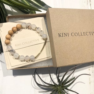 May include: A white and brown beaded bracelet with a gold bar accent, packaged in a brown box with the text "KINI COLLECTIVE" printed on it.