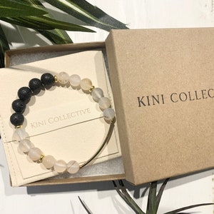 May include: A black lava rock and white agate beaded bracelet with a gold bar clasp. The bracelet is packaged in a brown box with the text "KINI COLLECTIVE" printed on the top.
