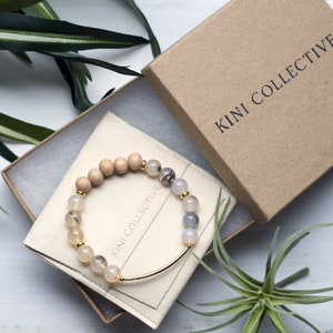 May include: A wooden and agate beaded bracelet with a gold bar clasp. The bracelet is packaged in a brown box with the text "KINI COLLECTIVE" printed on the top.