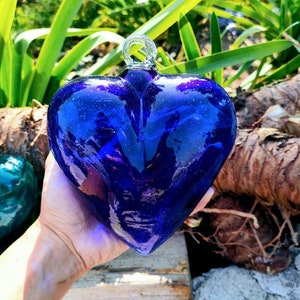Puede incluir: Un adorno de corazón de vidrio azul oscuro grande con una superficie texturizada. El corazón cuelga de un lazo plateado.