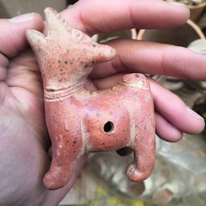 Pode incluir: Pequena figura de cerâmica castanho-avermelhada de um animal, possivelmente um cão ou um lobo, com um buraco na lateral. A figura tem uma cabeça detalhada com orelhas pontudas e uma cauda levantada. A superfície é salpicada com pontos mais escuros.