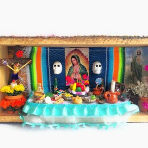 Puede incluir: Un altar miniatura del Día de Muertos con un mantel colorido, flores, velas y una imagen religiosa de la Virgen María. El altar está decorado con calaveras de azúcar, fruta y otras ofrendas tradicionales.