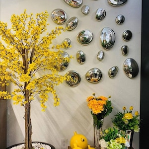 May include: A white wall with a collection of silver, round, reflective wall decor. A yellow flowering tree is in the left corner of the image. There are three yellow ceramic bird figurines and a vase with yellow flowers on a table in the bottom right corner.