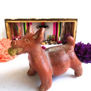 Pode incluir: Uma figurinha de cão de terracota com uma maçã verde na boca. O cão está em pé em frente a um pequeno santuário de madeira com figuras religiosas e velas dentro.