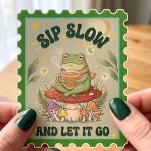 May include: A rectangular sticker with a green scalloped border featuring a green frog wearing a scarf, sitting on a large mushroom while holding a steaming mug. The text reads SIP SLOW AND LET IT GO, surrounded by small flowers, leaves, and stars.