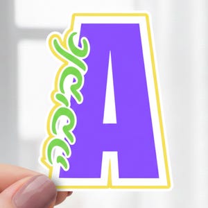 Puede incluir: Una pegatina con la letra « A » morada, una línea blanca en el centro, bordeada de amarillo y blanco. La palabra « Jerse » está escrita en verde en el lado izquierdo. La pegatina está sujeta por una mano.