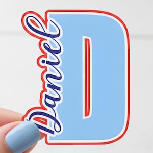 May include: A light blue sticker in the shape of the letter "D" with the name "Daniel" in cursive script. The letter and name are outlined in red and blue.