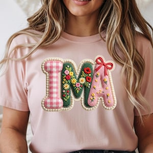 Könnte beinhalten: Hellrosa T-Shirt mit dem gestickten Wort "IMA". Das "I" hat ein rosa-weißes Vichy-Muster, das "M" ist grün mit Blumenstickerei und das "A" ist rosa mit einer roten Schleife und Blumenstickerei.