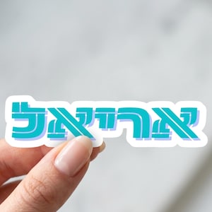 May include: A teal and white sticker featuring the Hebrew word "אריאל". The sticker is held by a hand with a light skin tone. The background is a soft, blurred white.
