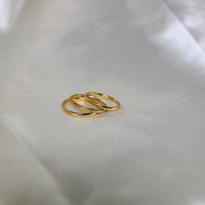 May include: Three delicate gold rings are displayed on a white, silky fabric. The rings are thin and have a polished finish, with one featuring a textured band. The rings are likely for the fingers and are a simple, elegant jewelry set.