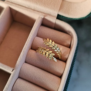 May include: Two gold-colored leaf-shaped rings rest inside a pink jewelry box. The rings have a delicate, detailed design, with each leaf vein visible. The jewelry box is open, revealing a soft, plush interior.