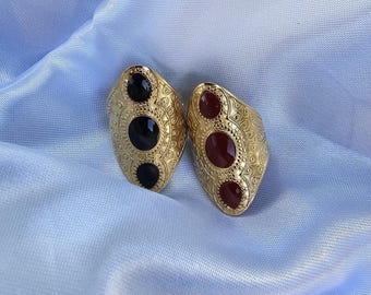 May include: Two gold-colored rings with oval-shaped faces. One ring features two black stones and one black stone, while the other has two red stones and one red stone. The rings have a textured, ornate design.