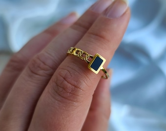 May include: A gold-colored ring with a chain-link band and a rectangular black stone. The ring is displayed on a finger against a soft, light blue background. The ring's design is modern and elegant.