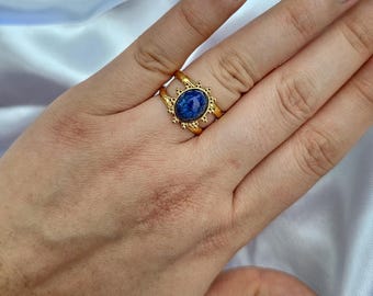 May include: A gold-colored ring with a large, oval, deep blue stone. The ring has a detailed, sunburst-like design around the stone and on the band. The ring is on a finger, showcasing the jewelry.