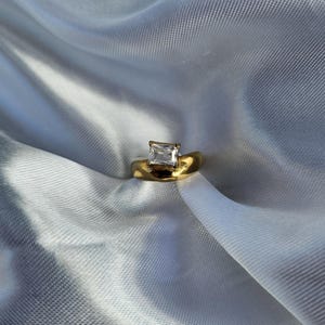 May include: A gold ring with a square, clear gemstone set on a wide band. The ring is displayed on a white, textured fabric, creating a contrast between the shiny metal and the soft background. The ring's design is simple yet elegant.