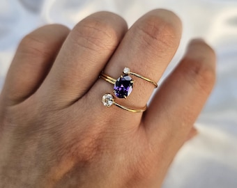 May include: A gold-colored ring with a large, oval, deep purple gemstone and two smaller clear gemstones. The ring is worn on a finger, with the gemstones positioned on either side of the finger. The ring's design is minimalist and elegant.