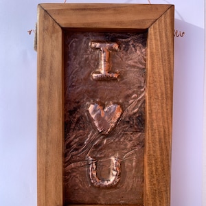 May include: A rectangular wall hanging featuring a wooden frame surrounding a textured copper panel. The copper centre displays the embossed letters I and U with a heart shape in the middle, all finished in a metallic copper tone. A thin copper wire loop is attached to the top for hanging.