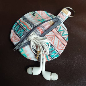 May include: A circular earbud case with a zip, containing white earbuds. The case has a colourful geometric pattern in coral, teal, and grey. A small loop with a silver key ring is attached. The earbuds are white with a gold-coloured jack.