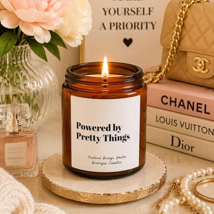 May include: A lit candle in a brown glass jar with a white label that reads "Powered by Pretty Things" and "Fashion Brings Smiles Boutique Candles." The candle sits on a round, white stone coaster with a gold-painted, textured edge.