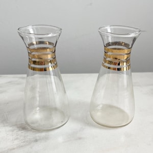 May include: Two clear glass bottles with gold bands around the middle. The bottles are on a white surface.