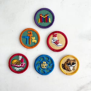 Puede incluir: Una colección de seis parches bordados circulares con varios diseños: una pila de libros coloridos, un juego de herramientas azul y naranja, una zapatilla blanca con rayas rojas, un gato amarillo sobre una alfombra rosa, un símbolo de reciclaje azul y un mapache marrón.