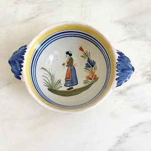 May include: A round ceramic bowl with two blue handles, featuring a hand-painted illustration of a person in a blue skirt and orange apron standing in a field. The interior is decorated with yellow and blue circular bands and floral motifs.