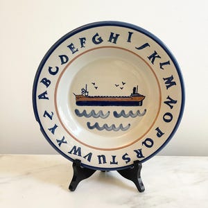 May include: A round ceramic plate with a cream-colored center featuring a hand-painted brown and blue cargo ship sailing on stylized blue waves with small birds above. The rim is decorated with the alphabet in dark blue capital letters.