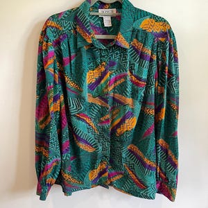 Könnte beinhalten: Ein langärmeliges Hemd mit Knopfleiste und einem lebendigen abstrakten Muster in den Farben Petrol, Magenta, Orange und Violett. Das Kleidungsstück hat einen klassischen Kragen, geraffte Schultern und geknöpfte Manschetten mit einem Grafikdruck.