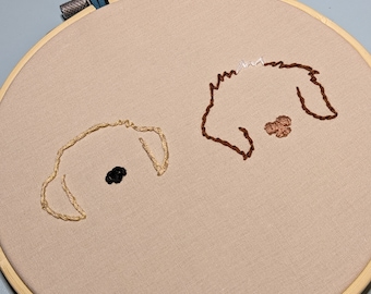 May include: Embroidery hoop featuring two dog faces stitched with thread. One dog is light tan with a black nose, the other is brown with a pink nose. The hoop is made of wood and has a blue fabric loop.