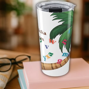 Pode incluir: Caneca de viagem branca com tampa preta, com um desenho de palmeira verde e letras coloridas. A caneca é cilíndrica e está sobre uma pilha de livros. A caneca tem cerca de 20 cm de altura.