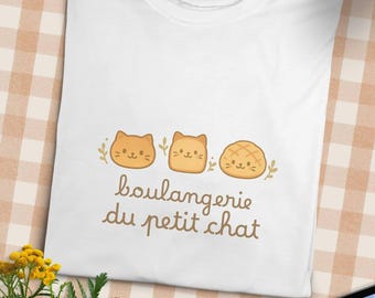 May include: White t-shirt featuring a design with three cartoon cat faces and the text "boulangerie du petit chat" in brown script. The cat faces are in shades of orange and brown, with green leaf accents.