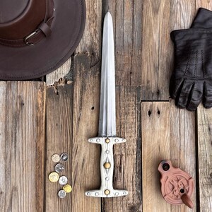 Può includere: Un pugnale bianco e argento con manico in legno e dettagli dorati. Il pugnale è appoggiato su una superficie in legno con un cappello di pelle marrone e un paio di guanti di pelle marrone.