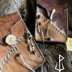 May include: Two handmade leather pouches with decorative stitching and wooden bead accents. The pouches are various shades of brown, with a natural, rustic aesthetic. One pouch features a bone accent and a wooden button closure.