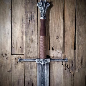 May include: A medieval-style sword with a silver blade and guard, a brown leather-wrapped handle, and a decorative silver pommel. The sword is displayed against a rustic wooden background, showcasing its intricate design and craftsmanship.