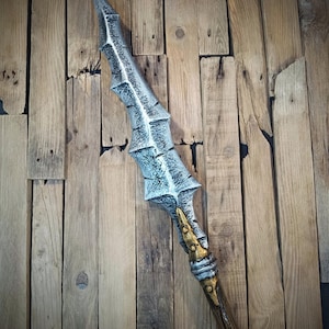 May include: A spear with a long, jagged, silver-coloured metal blade featuring a textured, weathered finish. The base of the blade connects to a brown, wood-textured shaft with gold-coloured accents and silver-coloured rings at the transition point.