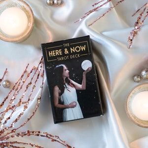 May include: A black tarot deck box with gold lettering that reads "The Here & Now Tarot Deck". The box is on a white satin surface with gold and silver decorative accents.