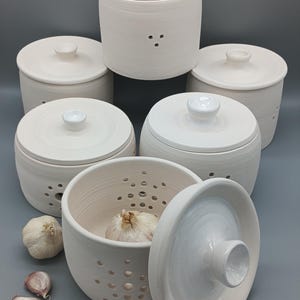 Op de afbeelding: Een verzameling van zes cilindervormige keramische knoflookpotten met bijpassende deksels. Elke pot heeft een matte gebroken witte afwerking en kleine ventilatiegaatjes die in cirkelvormige patronen aan de zijkanten zijn aangebracht.