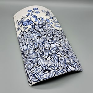 Può includere: Un appendiabiti da parete in ceramica bianca con un design floreale blu e nero. I fiori sono stilizzati e hanno un aspetto fantasioso. L'appendiabiti da parete è curvo e ha un foro nella parte superiore per appenderlo.