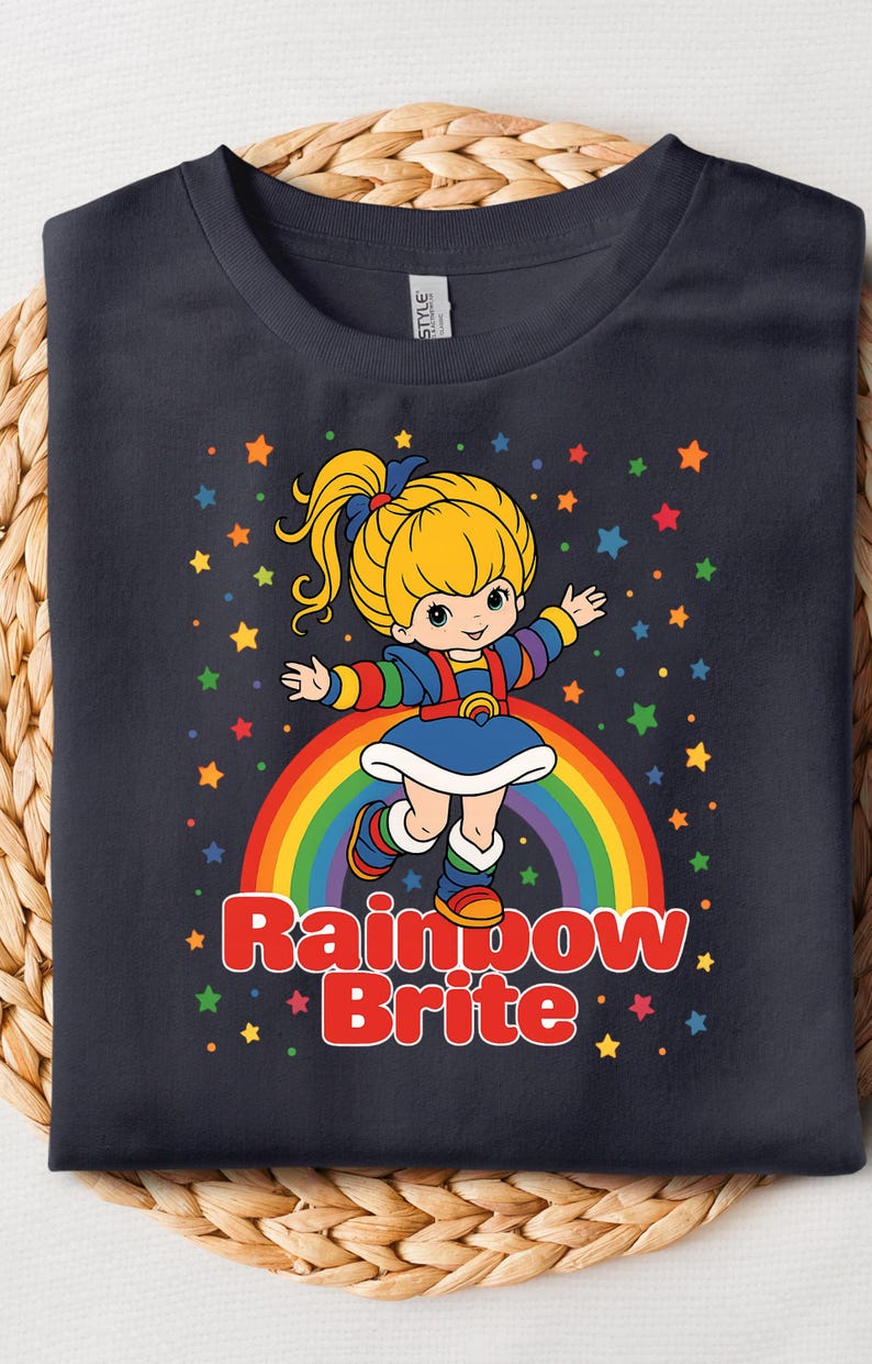 May include: Navy blue t-shirt featuring a cartoon image of Rainbow Brite standing on a rainbow. The design includes colorful stars and the words "Rainbow Brite" in red. The shirt is displayed on a woven surface.