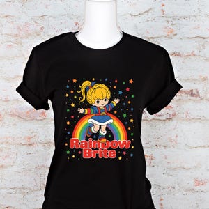May include: Black t-shirt featuring a colorful graphic of a blonde character on a rainbow with the text "Rainbow Brite." The design includes stars and a cheerful aesthetic. The shirt is displayed on a mannequin.