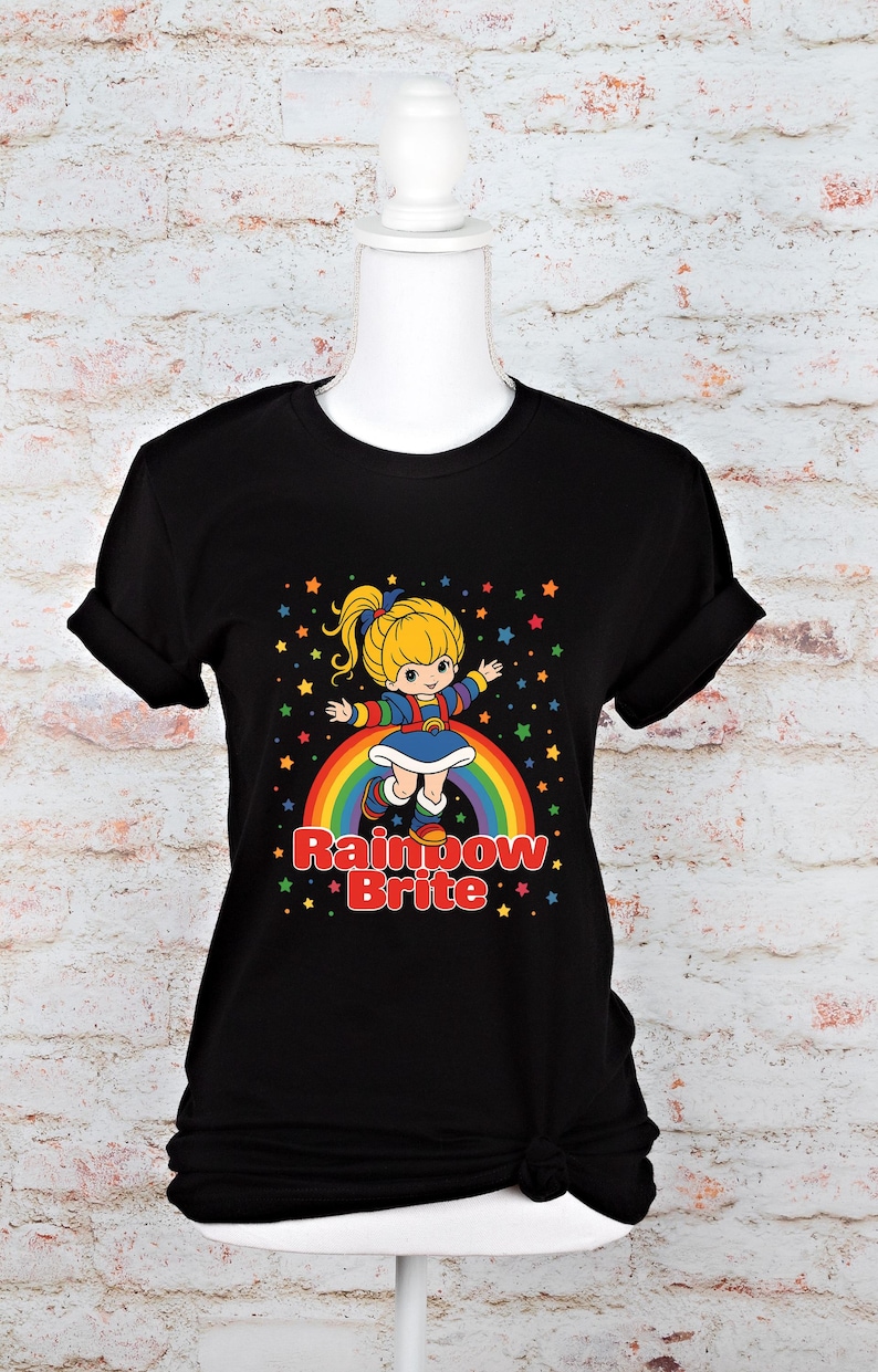 May include: Black t-shirt featuring a colorful graphic of a blonde character on a rainbow with the text "Rainbow Brite." The design includes stars and a cheerful aesthetic. The shirt is displayed on a mannequin.