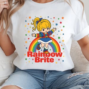May include: White t-shirt featuring a colorful graphic of Rainbow Brite standing on a rainbow. The design includes stars and the text "Rainbow Brite" in red. The shirt is a classic crew neck.